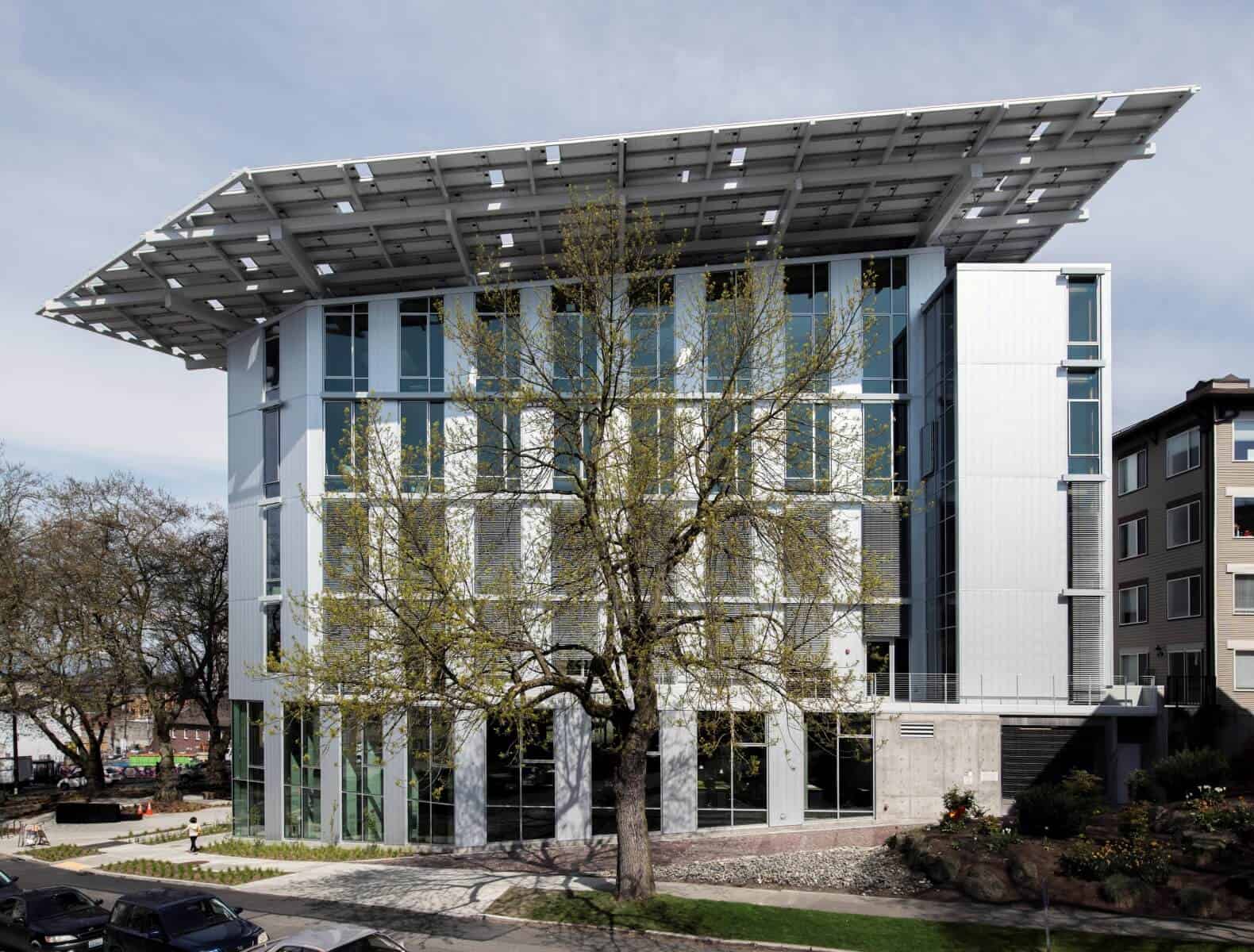 A view of the Bullitt Center, a modern and sustainable office building with a distinctive large overhanging roof, glass facade, and solar panels, located in an urban setting with surrounding trees and residential buildings.