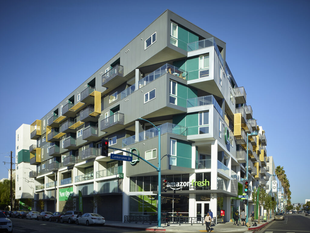 Modern mixed-use building with Amazon Fresh store on the ground level, featuring multiple balconies, colorful panels, and situated on a busy street corner.