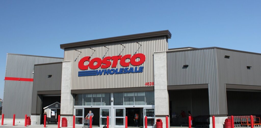 A modern Costco Wholesale building with a prominent red and blue logo on its gray metal facade, featuring large glass entrance doors and a few customers with shopping carts, set against a clear blue sky.