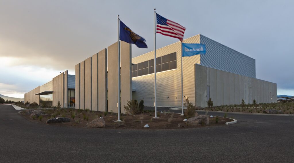 Facebook Prineville Data Center, a modern industrial building with a sleek metal exterior and vertical concrete panels, situated under a cloudy sky. The facility is fronted by three flagpoles displaying the Oregon state flag, the American flag, and the Facebook flag, with neatly landscaped surroundings.