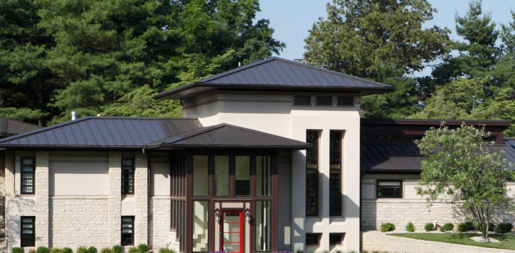 Modern residence with a dark metal roof and beige walls, surrounded by lush greenery and trees.