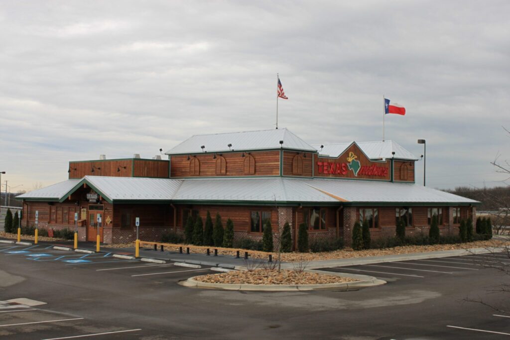 Texas Roadhouse restaurant building with a rustic design, wooden exterior, green trim, and metal roofing, accompanied by American and Texas flags.